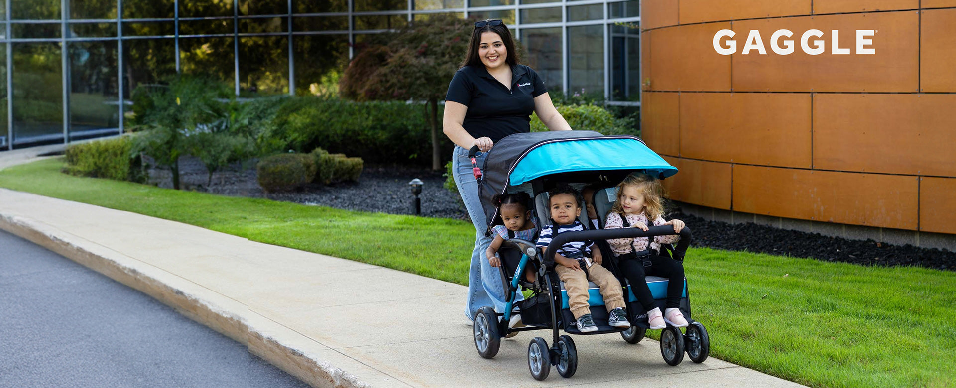 A woman pushes a four-child stroller with a bright turquoise canopy on a sidewalk next to a modern building.