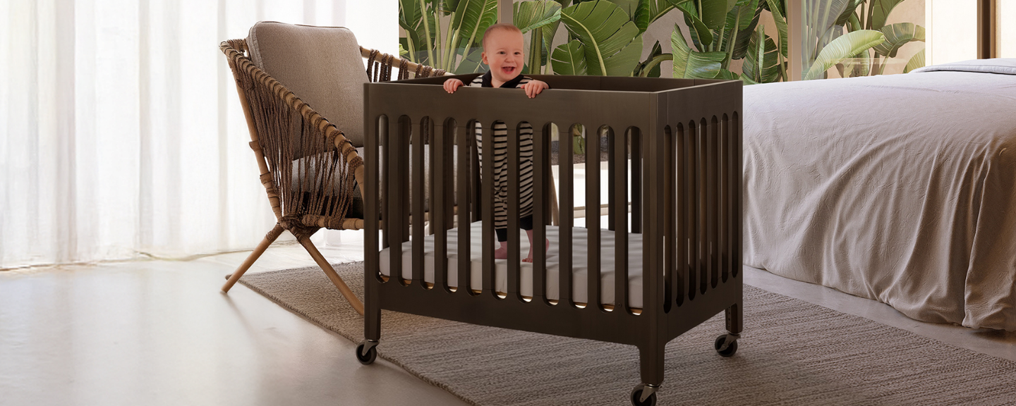 Child playing in a wooden crib in a bedroom with a bed and chair in the background.